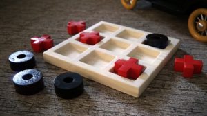 White tray with nine sections. Large red x shaped pieces and black circle pieces are in or near this Tic Tack Toe game tray.