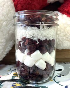 Fruit and nut snack mix layered in a small clear jar. Cherries, marshmallows, cranberries, and coconut. This layering resembles a candy cane.