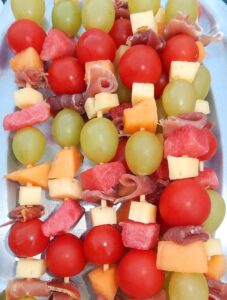 Plate of mixed food on skewers, such as grapes, melon, tomatoes, cheese, and salami.