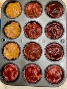12 cup muffin pan filled with raw meatloaf mixture with 4 different toppings including ketchup, BBQ sauce, salsa and honey mustard glaze.