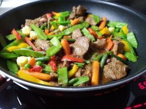 Skillet of stir-frying carrots, yellow bell pepper, snow peas, green beans, beef and sauce.