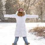 Woman in winter coat , arms stretched out and smiling face looking up to the falling snow.