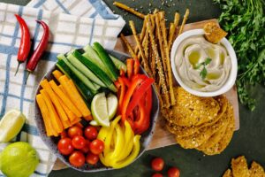 A cutting board topped with a bowl of cut fresh vegetable (carrots, cucumber, red bell peppers, yellow bell peppers, cherry tomatoes and lime wedges), bread sticks, flat crackers and a bowl of hummus dip.