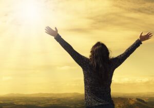 Person standing with arms wide open and eyes to the sky, while facing the sunrise.