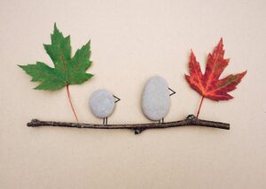 2 rocks glued to a paper with a beak and legs added to resemble birds. They are on a stick with a maple leaf at each end of the stick.