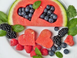 A slice of watermelon with heart shapes cut out and filed in with fresh blueberries. The watermelon heart shapes are surrounded by fresh blackberries, blueberries, raspberries and mint leaves. 