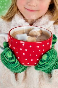 Child with green mittens holding a large red and white mug of steaming hot chocolate with 3 marshmallows